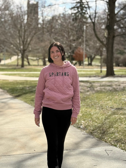 Woman standing outdoors wearing a quilted pink sweatshirt with the word "Spartans" embroidered on the front. In each letter of "Spartans" is a floral pattern. The sweatshirt has a middle pocket and drawstrings. 