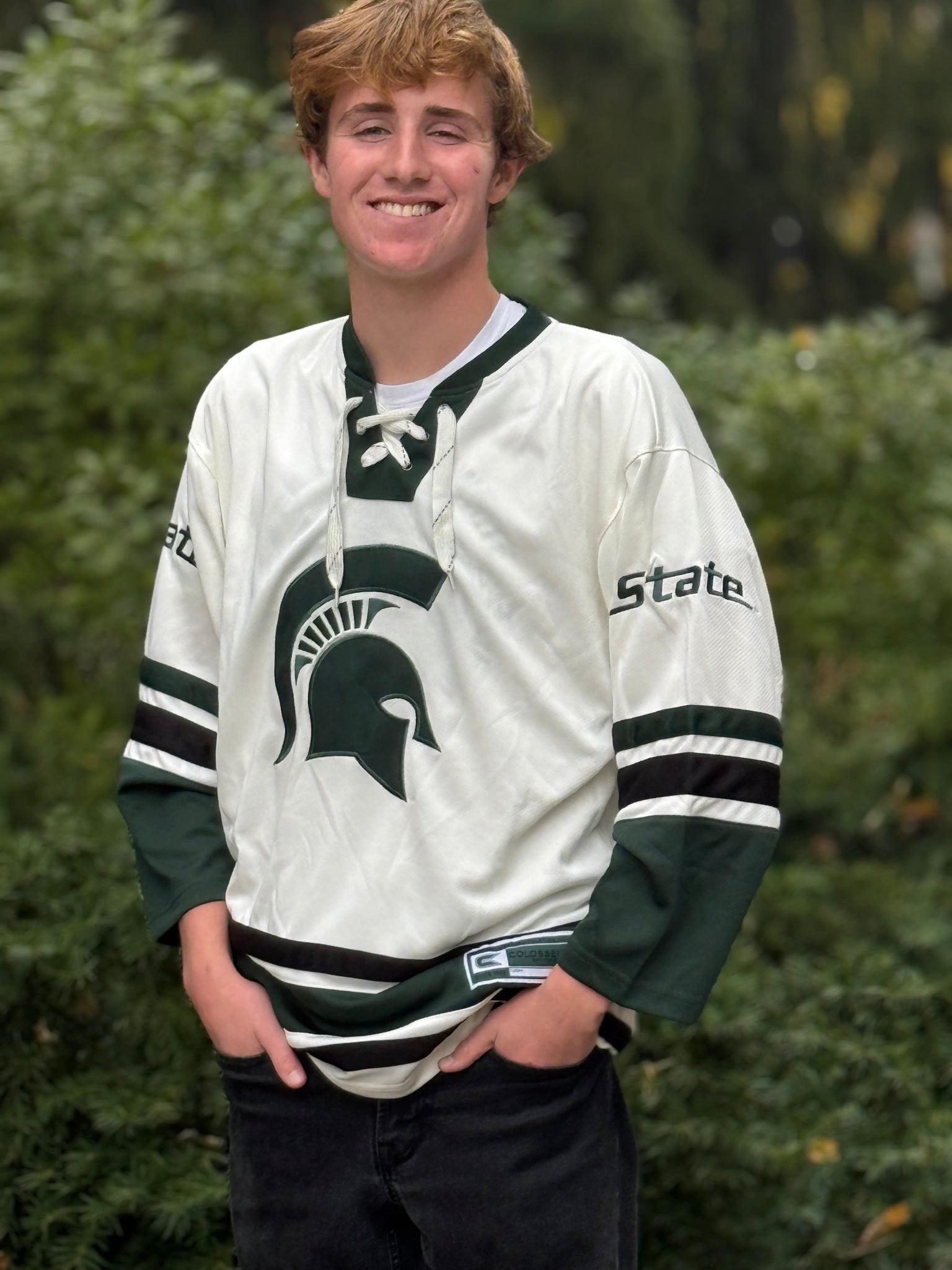 A person wearing an off white hockey jersey with green and black accents, featuring a green Spartan helmet logo on the front, green 'State' text on both sleeves, striped sleeves,  and  lace-up collar.