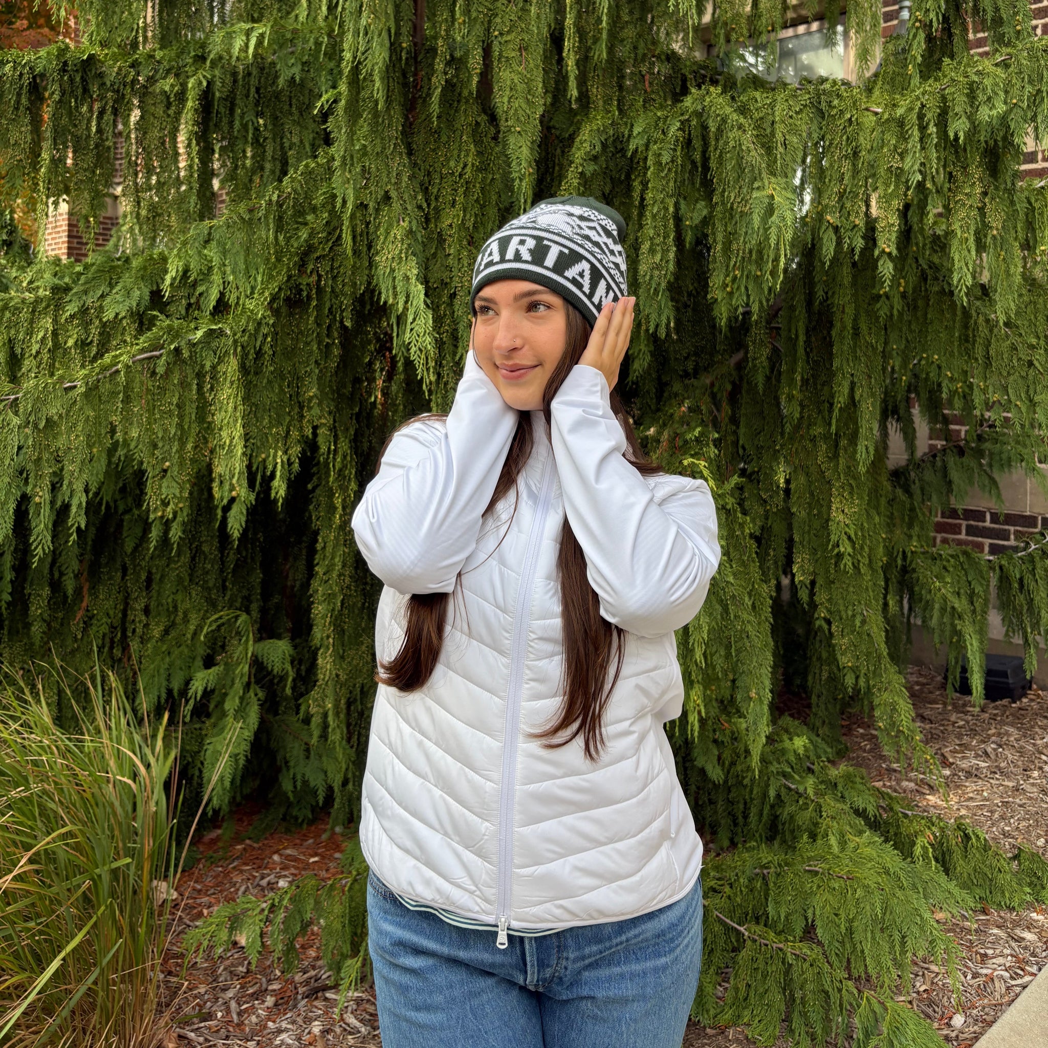 Woman standing outside, wearing a white two-way zip jacket with puff stitching on the middle section and a green Spartan helmet on the left chest. 