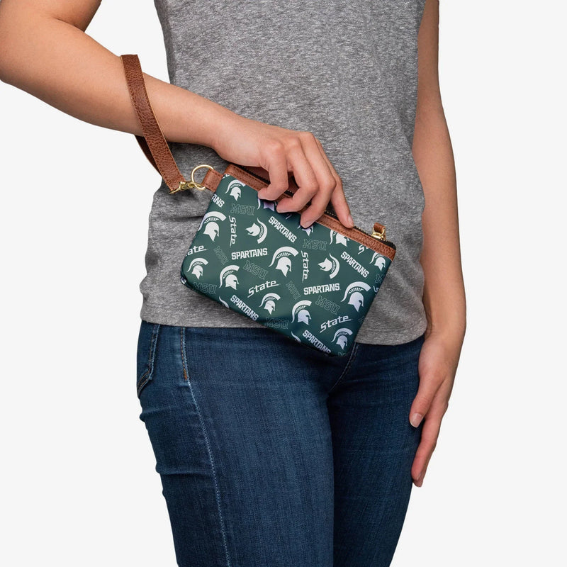 Person holding small rectangular green wristlet with white printed Spartans, State, and MSU work marks as well as white Spartan helmets all over design. Brown faux leather trim and wristlet loop. Zipper closure. 