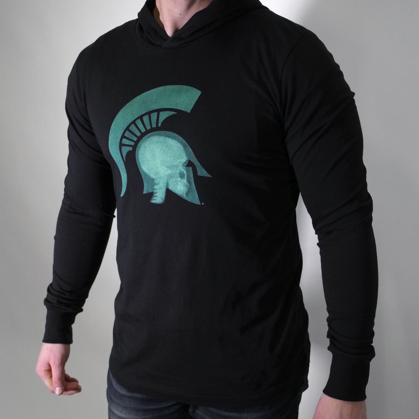 Front view of a man wearing a black long-sleeve crewneck T-shirt with a hood built-in. On the center chest is a graphic of an x-ray of a head wearing a Spartan Helmet.