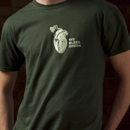 A person wearing a forest green short sleeve crewneck T-shirt with a diagram of a heart and text reading "We Bleed Green" printed in white on the center chest.
