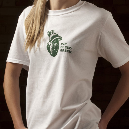 A person wearing a white short sleeve crewneck T-shirt with a diagram of a heart and text reading "We Bleed Green" printed in forest green on the center chest.