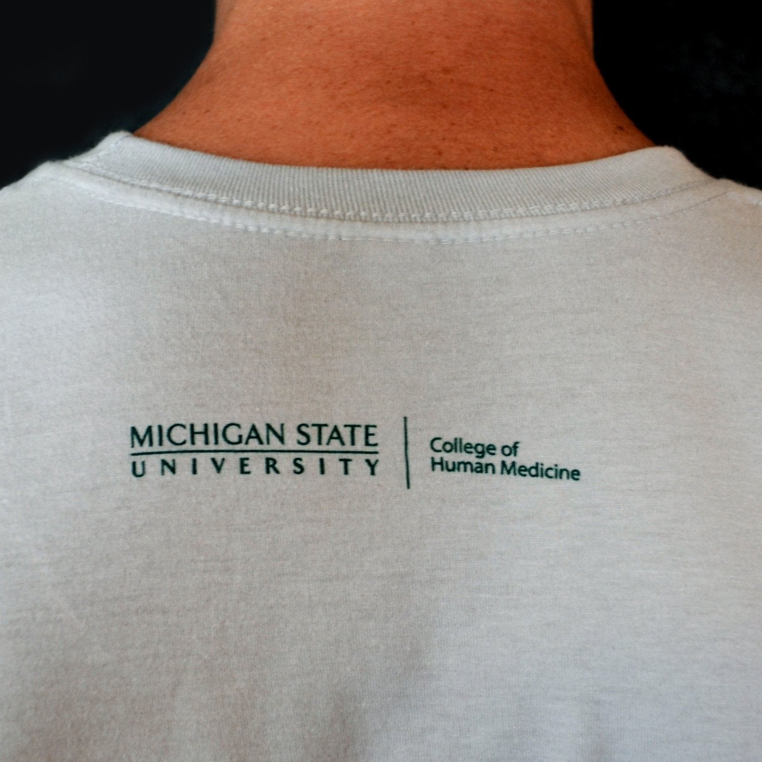 Close-up of the College of Human Medicine wordmark logo, printed in green just underneath the neckline on the back of the T-shirt.