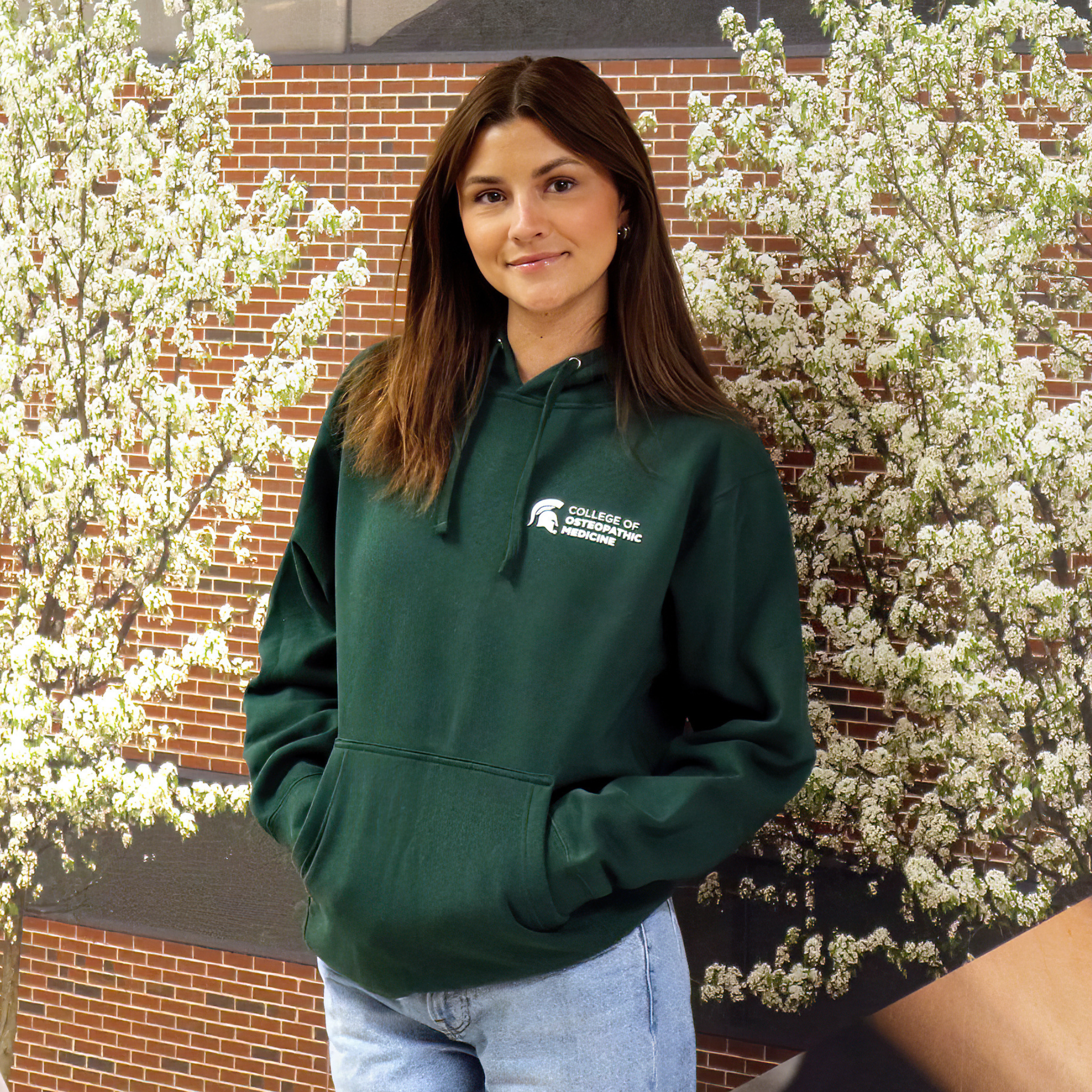 A person in a forest green hoodie sweatshirt. Features an adjustable drawstring hood, a front pocket, with College of Osteopathic Medicine logo embroidered in white on the left front side. 
