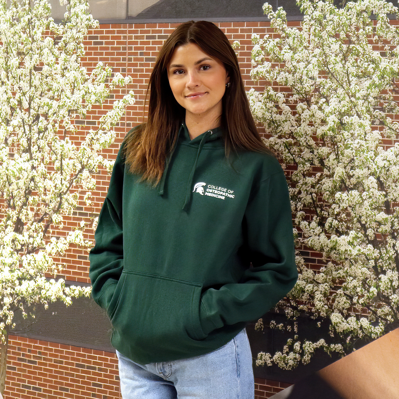 A person in a forest green hoodie sweatshirt. Features an adjustable drawstring hood, a front pocket, with College of Osteopathic Medicine logo embroidered in white on the left front side. 