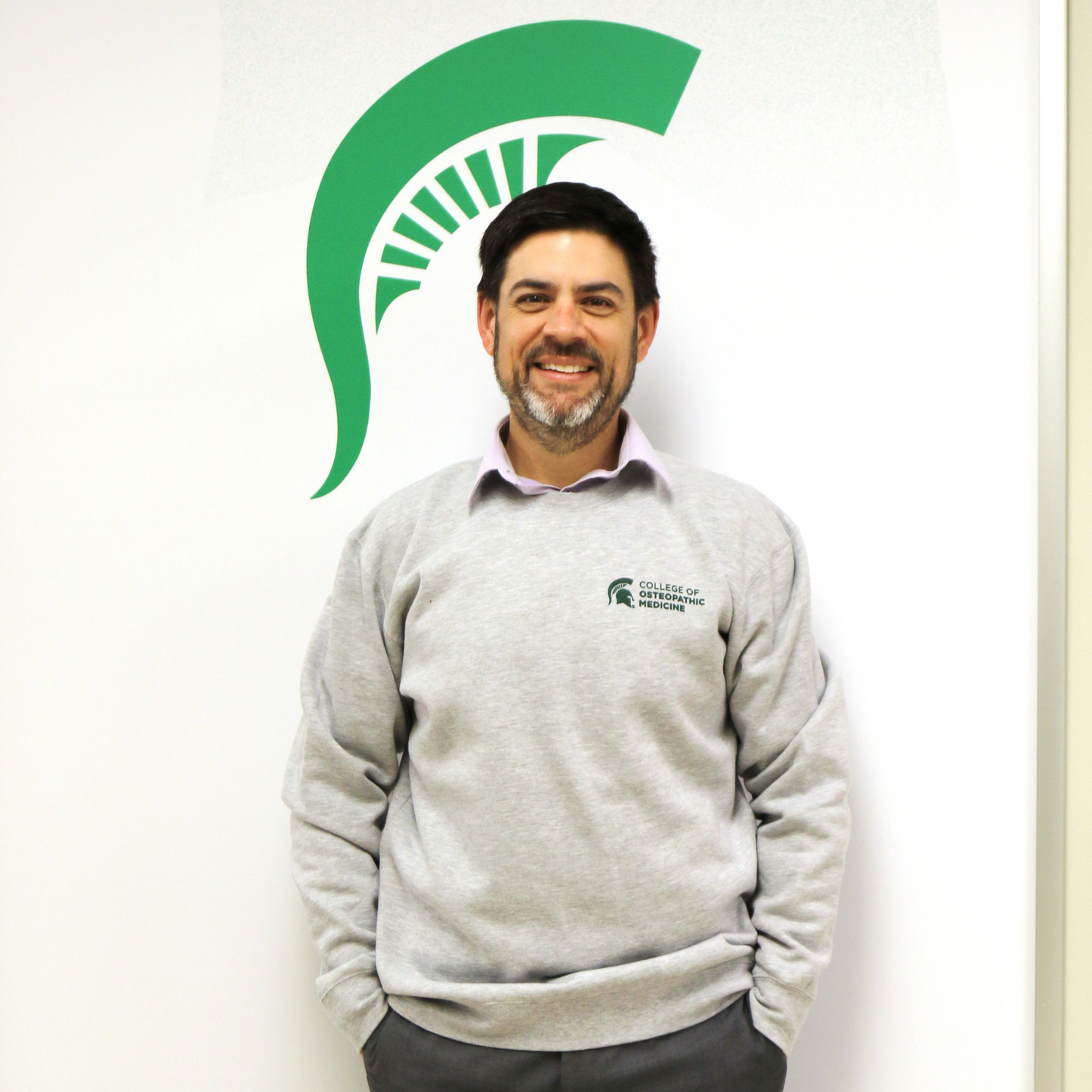 A person wearing a heather gray crewneck sweatshirt that features ribbed cuffs and a ribbed hem, with MSU College of Osteopathic Medicine printed in green on the left front.