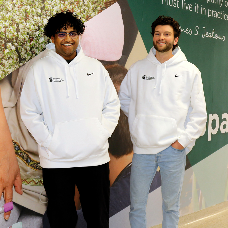 Two people with white Nike fleece hoodie sweatshirts featuring a green embroidered Nike Swoosh and green screen-printed MSU College of Osteopathic Medicine logo on the front.  