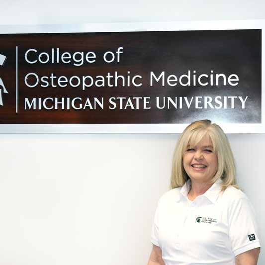 Person in a white fitted polo with forest green embroidered College of Osteopathic Medicine logo on the left upper chest. 