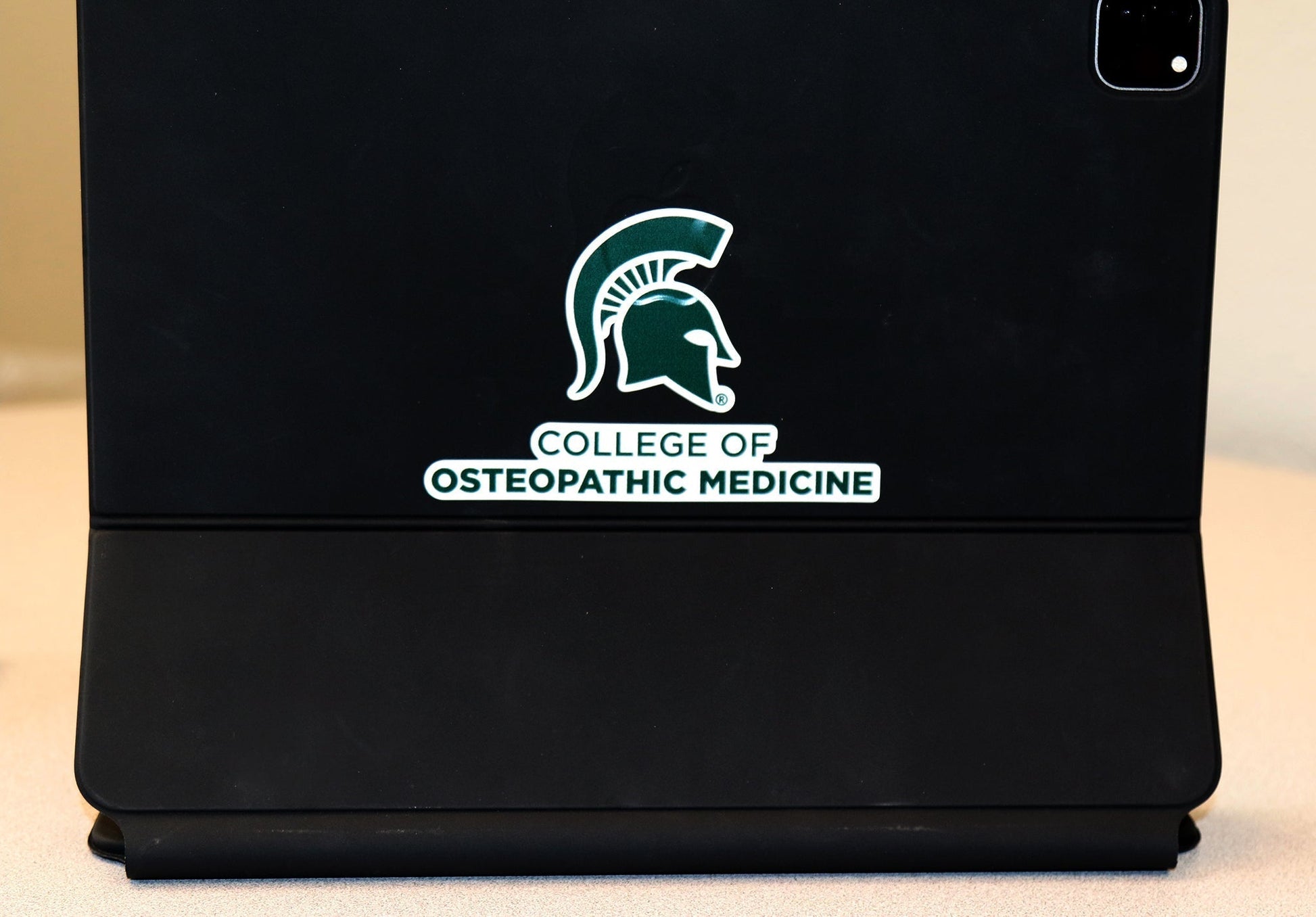 White vinyl transfer decal with a forest green College of Osteopathic Medicine logo and Spartan Helmet logo on it. Shown on top of a black iPad case. 