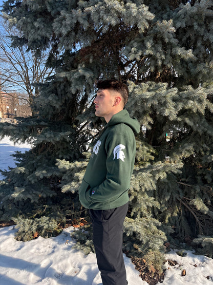 Man standing outdoors, showing the left side of a green hoodie. The left sleeve has a white Spartan Helmet logo. 