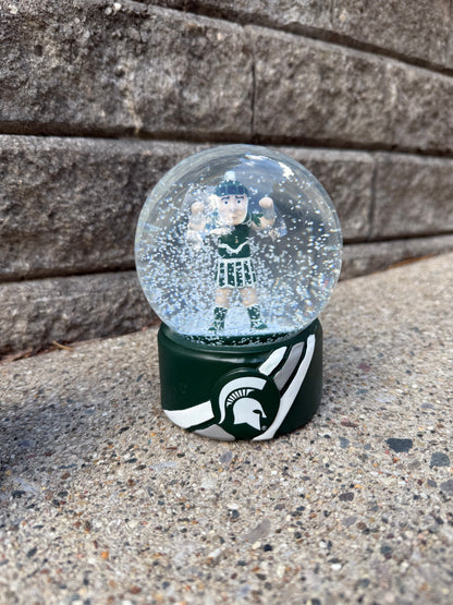 Sparty snow globe with a green base and Sparty figurine inside. The base has two white stripes, one gray stripe in the middle, and a Spartan helmet logo in the middle of the the stripes. 