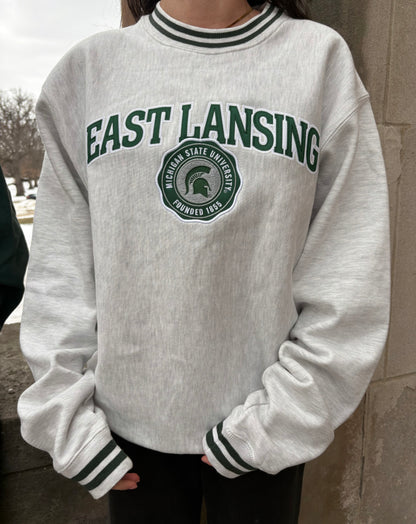 Close up of a white crewneck "East Lansing" written on the front chest and the Michigan State University seal logo with the Spartan Helmet logo inside, both in green and white. On the collar and sleeve cuffs are green and white stripes. 