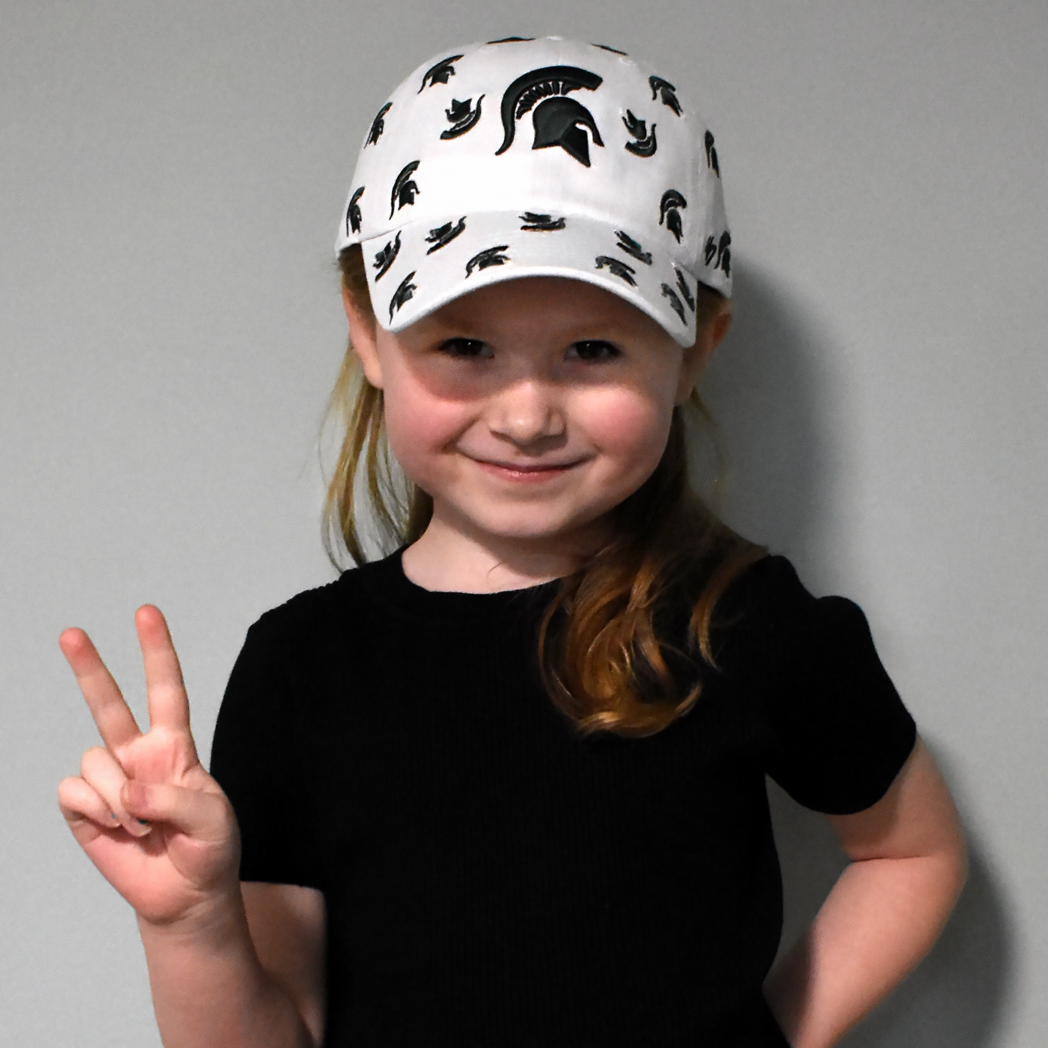 Child wearing a white cap with green Spartan helmet logos patterned all over it.