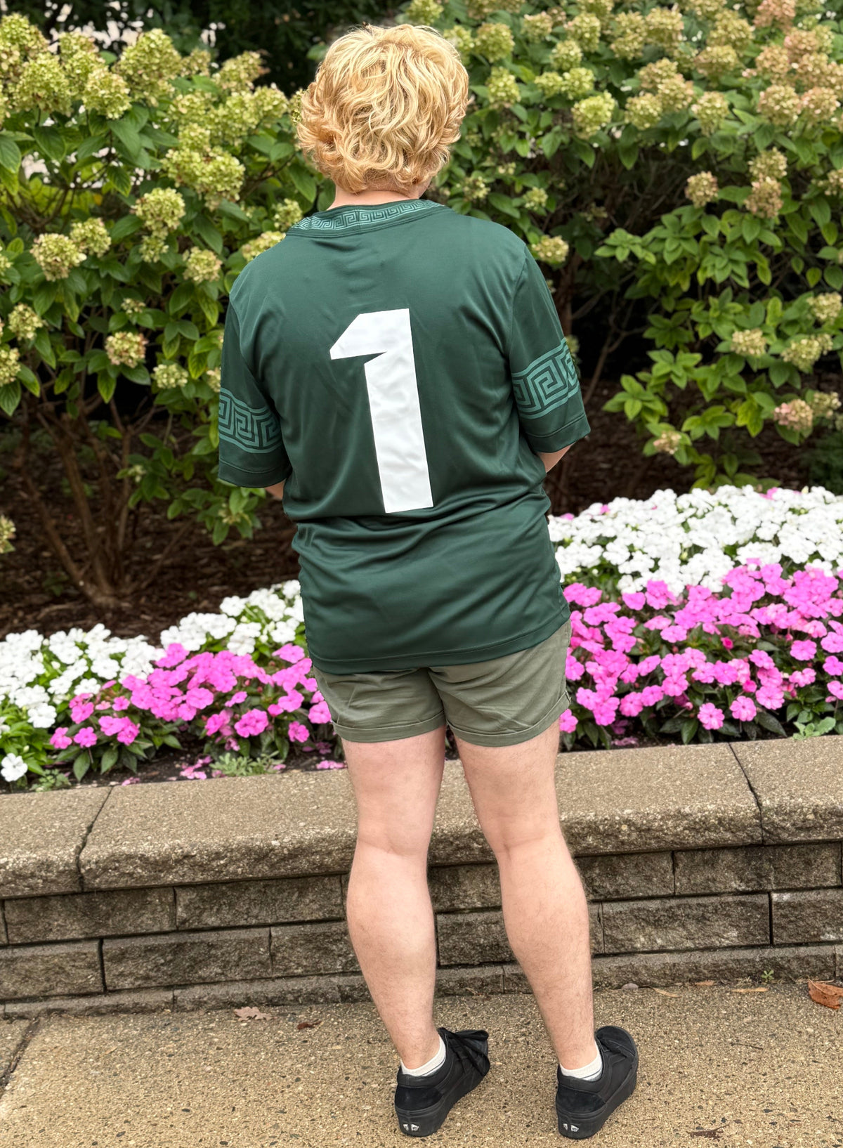 Back view of a person standing outdoors  wearing a green Michigan State Spartans football-style jersey. The back of the jersey features a large white number 1 centered on the dark green fabric, with geometric patterned sleeve accents and matching trim on the collar. The outfit is paired with olive green shorts, white socks, and black sneakers. A stone retaining wall and landscaped garden create the background in natural daylight.