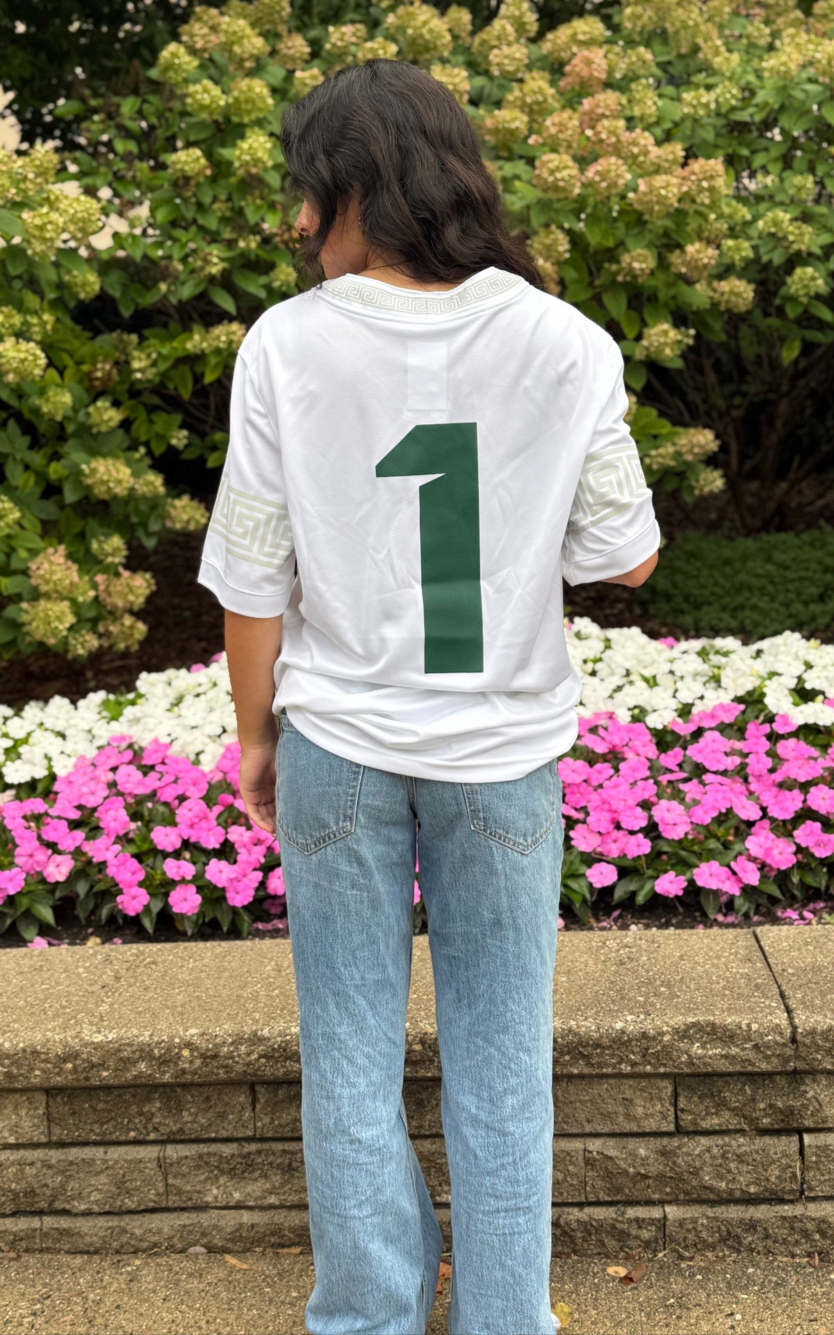 Back view of a person standing outdoors wearing a white Michigan State Spartans football-style jersey. The back of the jersey features a large green number 1 centered on the white fabric, with light gray geometric trim on the sleeves and collar. The outfit is paired with light-wash blue jeans. A stone retaining wall and landscaped garden provide the background in natural daylight.