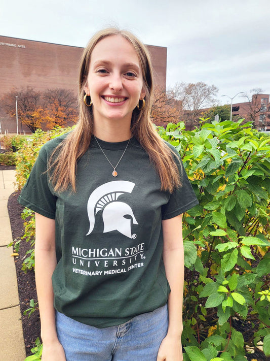 A woman in blue jeans wearing a green short sleeve T-shirt. Across the center chest is a large white Spartan helmet centered above the Michigan State wordmark logo. Beneath that is large block text reading “Veterinary Medical Center.”