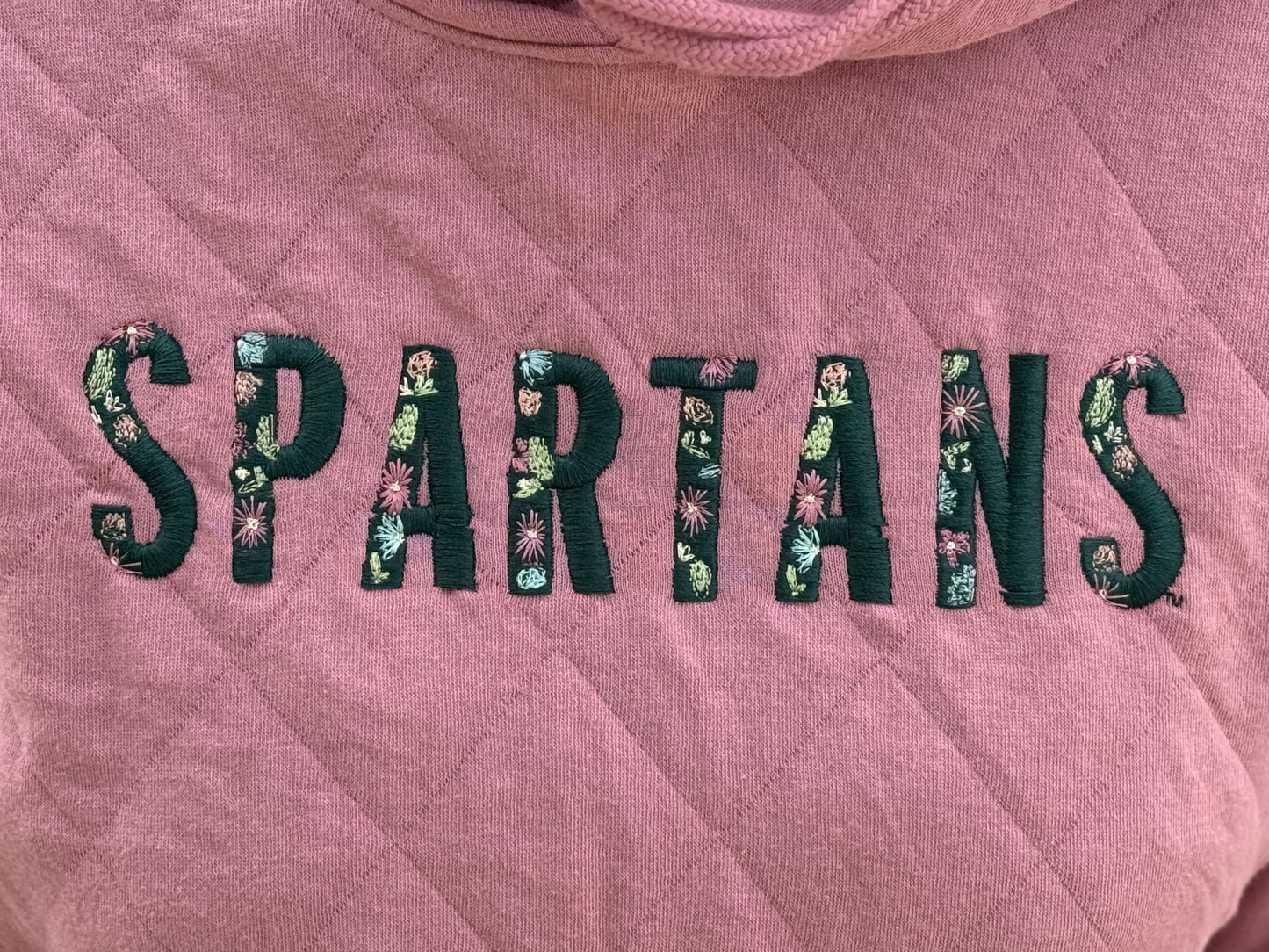 Close up of a quilted pink sweatshirt with the word "Spartans" embroidered on the front. In each letter of "Spartans" is a floral pattern.  