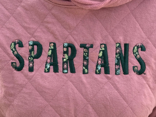 Close up of a quilted pink sweatshirt with the word "Spartans" embroidered on the front. In each letter of "Spartans" is a floral pattern.  