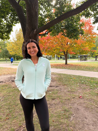 Woman standing outdoors, wearing a light blue striped full-zip jacket with a white embroidered Spartan Helmet logo on the left chest. 