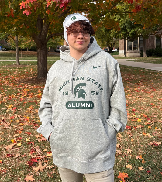 Person standing outdoors, wearing a gray Michigan State University Alumni drawstring hoodie with a curved "Michigan State" wordmark, the year "1855," the Spartan helmet logo, and a bold "Alumni" wordmark screen printed on the front in green. The Nike logo is also printed on the right upper chest in green. 