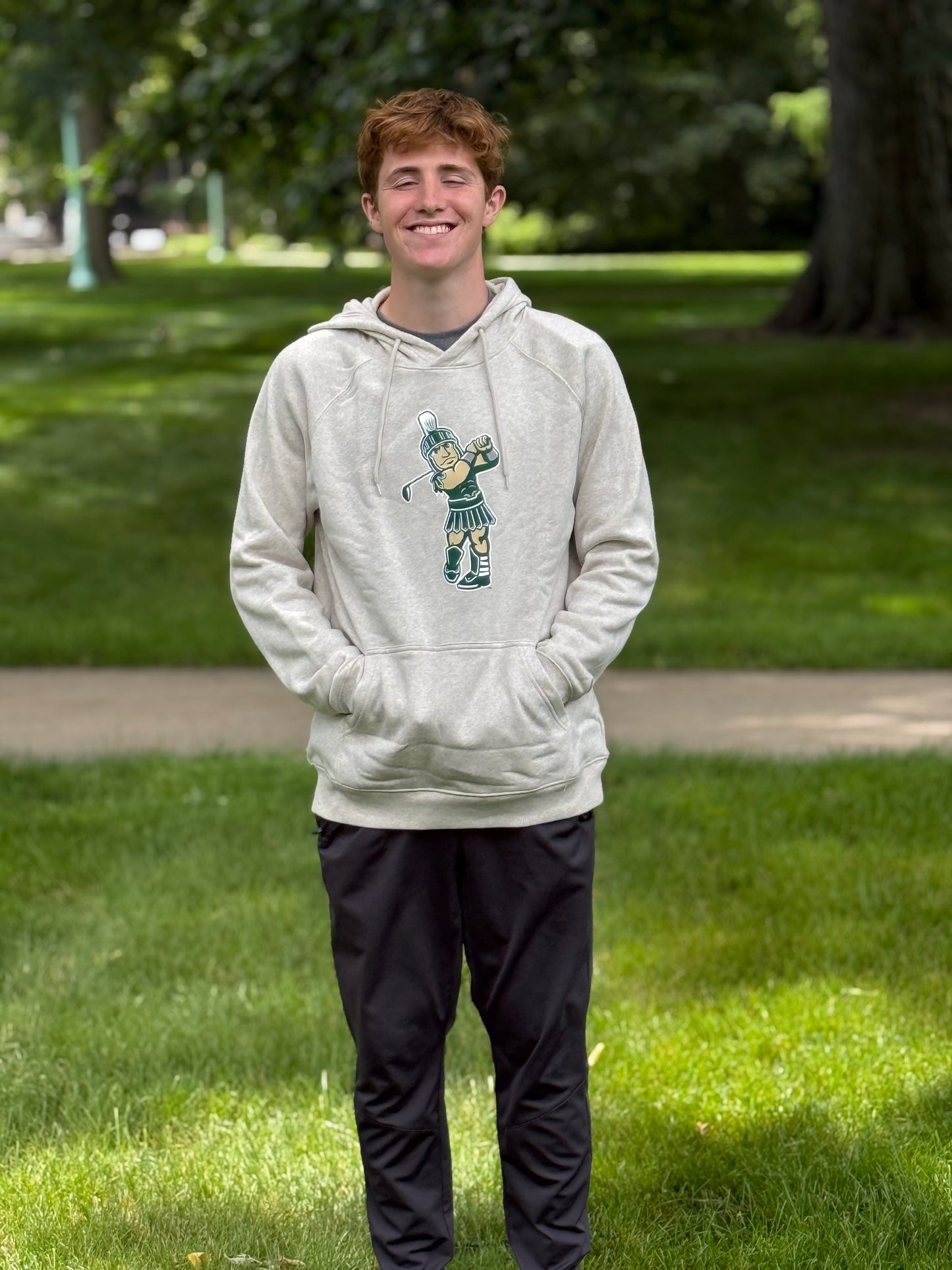Person modeling a heather oatmeal pullover hoodie featuring a Golf Sparty screen printed graphic on the chest, with a drawstring hood and front kangaroo pocket, paired with black athletic pants outdoors on a grassy campus setting.