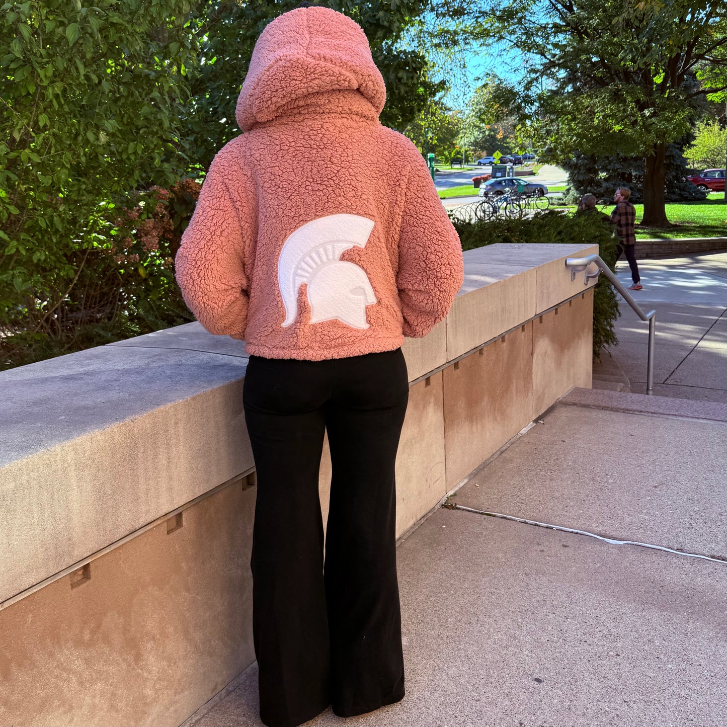 Woman standing outside, showing the back of a cropped pink lounger with a hood and large white Spartan Helmet logo centered on the back. 