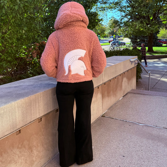 Woman standing outside, showing the back of a cropped pink lounger with a hood and large white Spartan Helmet logo centered on the back. 