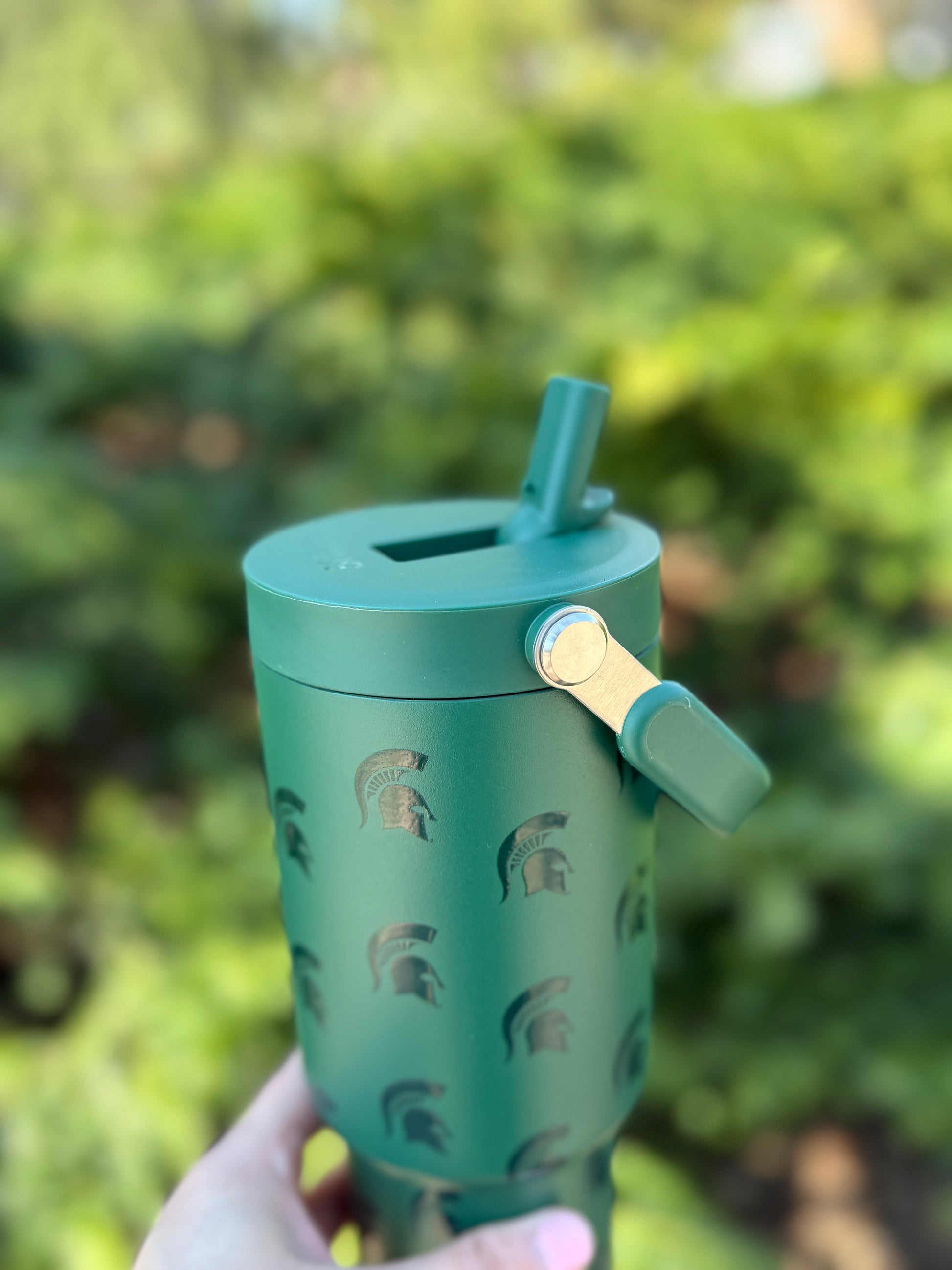 Close up of a green water bottle with a handle at the top, a straw that flips up and down, and dark green Spartan Helmet logos patterned all over. Shown outdoors. 