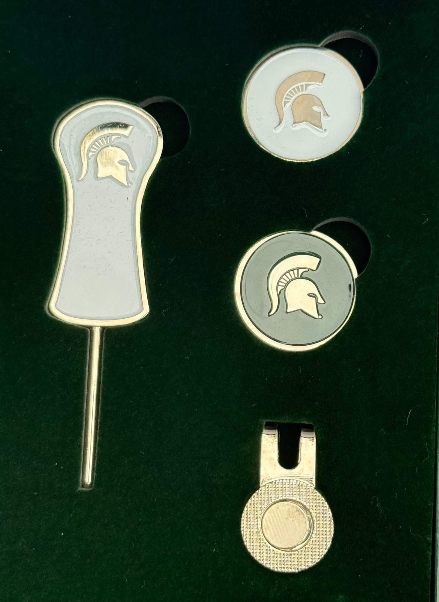 Divot tool that is silver outlined with white center and a silver helmet towards top of handle. Two round ball makers one is white with a silver Spartan helmet other is green with silver spartan helmet. Silver magnetic ball maker clip that clips on to bill of hat. 
