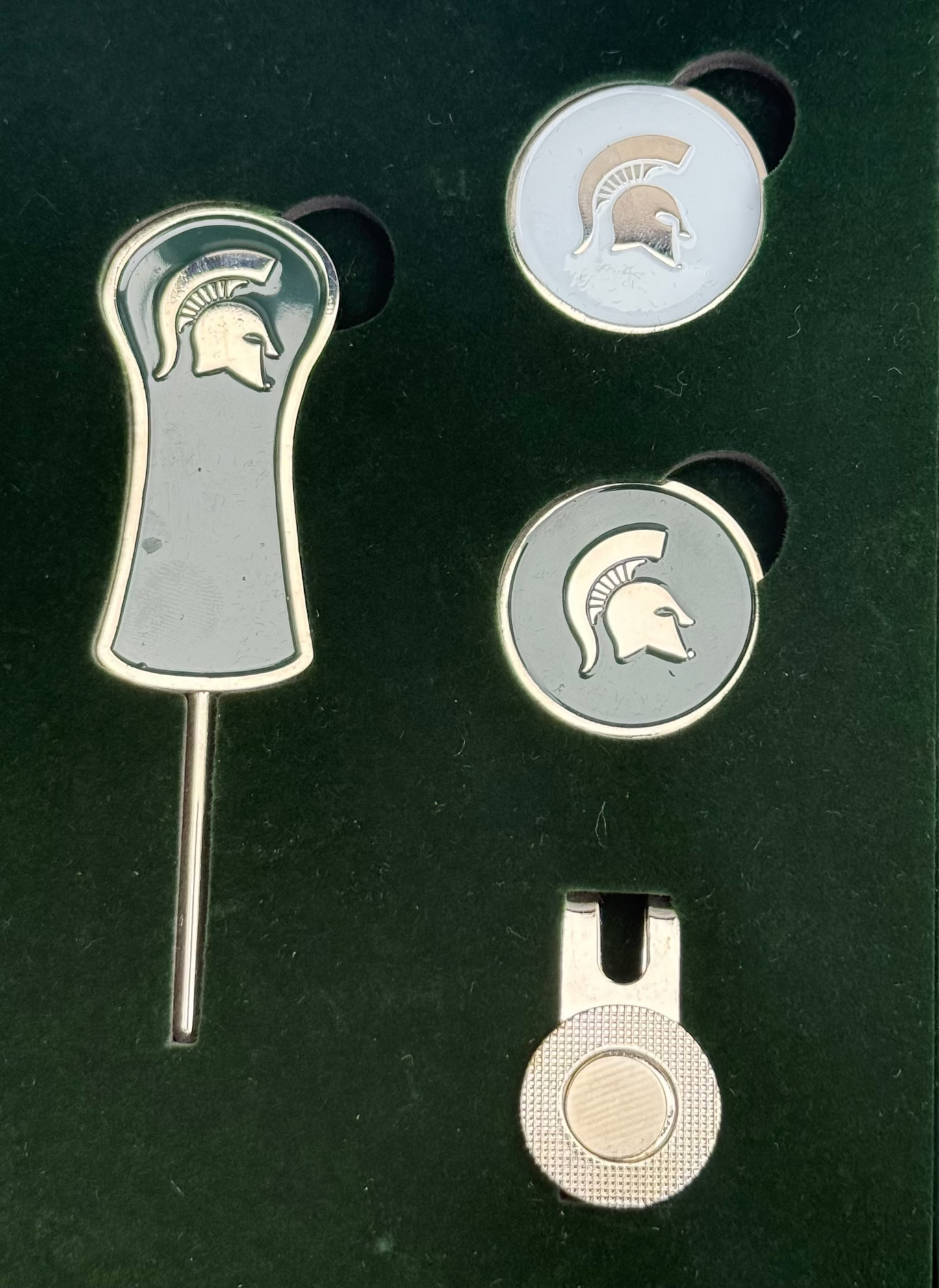 Divot tool that is silver outlined with green center and a silver helmet towards top of handle. Two round ball makers one is white with a silver Spartan helmet other is green with silver spartan helmet. Silver magnetic ball maker clip that clips on to bill of hat. 