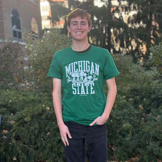 Person standing outdoors on a sidewalk covered with autumn leaves, wearing a green Michigan State University T-shirt with white text that reads “Michigan State” and features the Peanuts characters along with the phrases “Go Green!” and “Go White!” on either side of the characters. The person is also wearing black jeans, with one hand in a pocket. Background includes green bushes, tall trees, and part of a brick building.