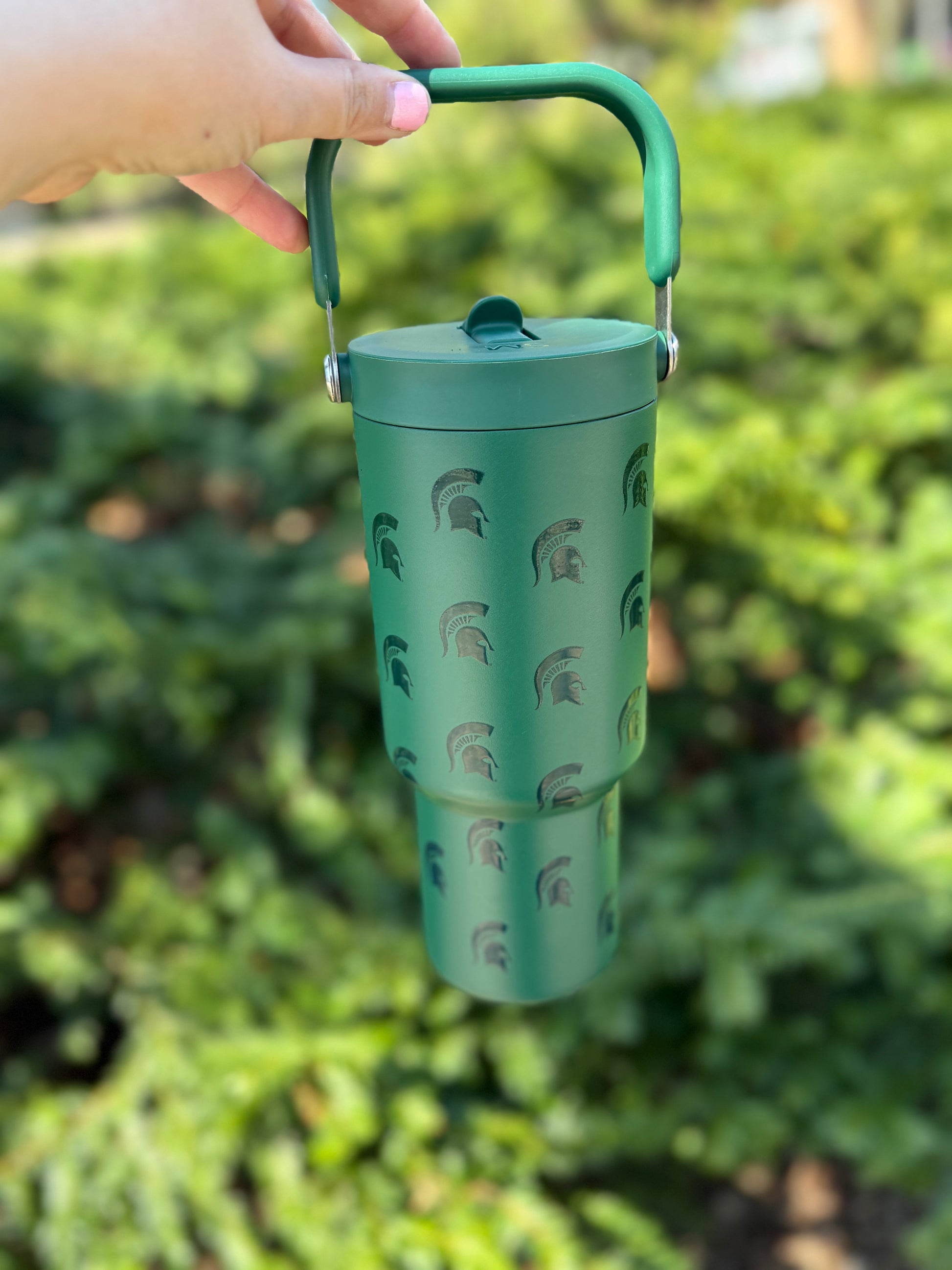 Green water bottle with a handle at the top, a straw that flips up and down, and dark green Spartan Helmet logos patterned all over. Shown outdoors. 