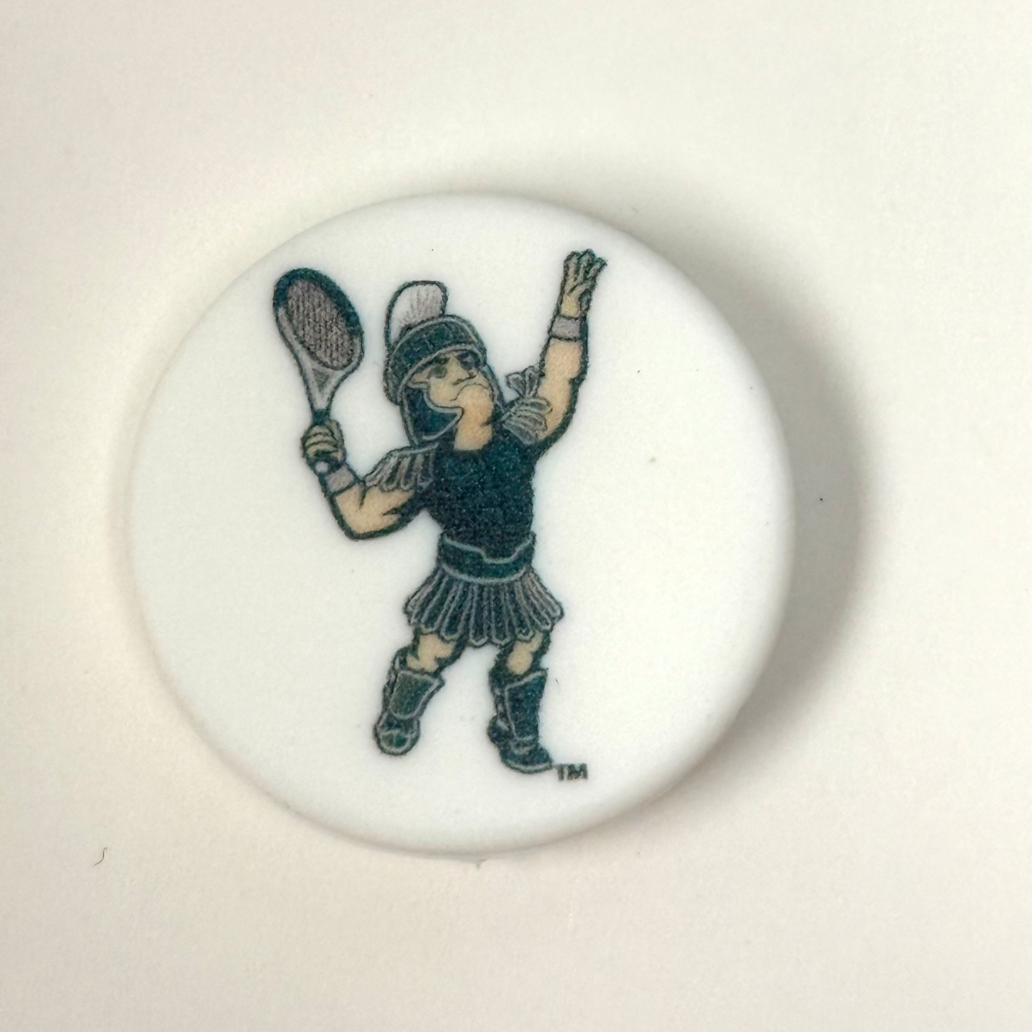 A white plastic round tennis dampener with Tennis Sparty centered on both sides. 