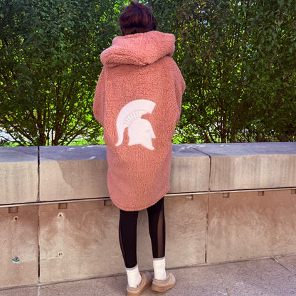 Woman standing outdoors, showing the back of a long pink lounger with a hood and white Spartan Helmet logo centered on the back. 
