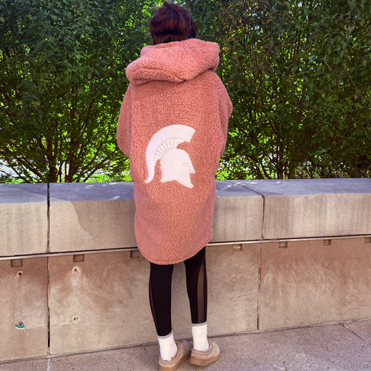Woman standing outdoors, showing the back of a long pink lounger with a hood and white Spartan Helmet logo centered on the back. 