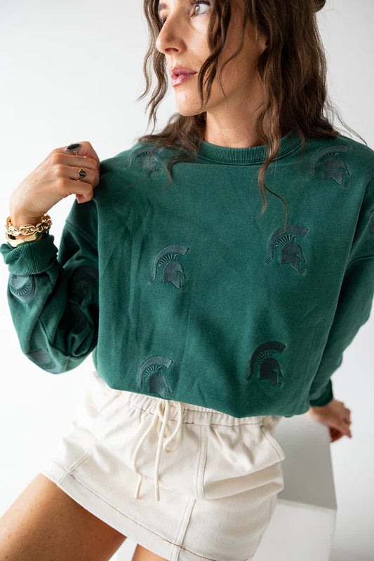 Woman modeling an oversized forest green crewneck sweatshirt featuring subtle tonal embroidered Spartan helmet designs scattered across the front. 