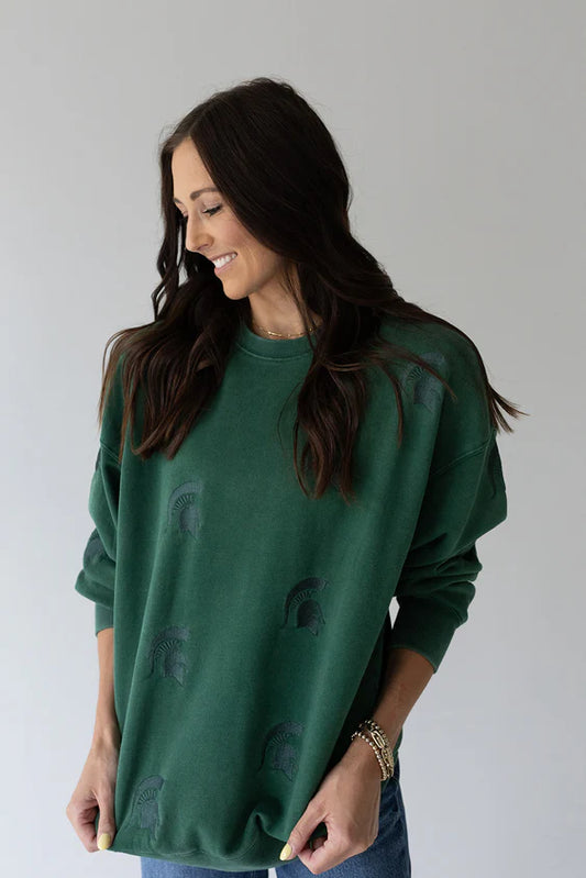 Woman modeling an oversized forest green crewneck sweatshirt featuring subtle tonal embroidered Spartan helmet designs scattered across the front. The relaxed-fit sweatshirt is paired with light-wash jeans and styled with rolled sleeves against a neutral background.
