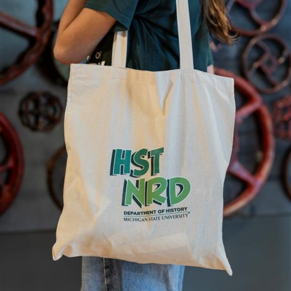 Person holding an off-white tote bag that reads "'HST NRD,' Department of History, Michigan State University" in green. 