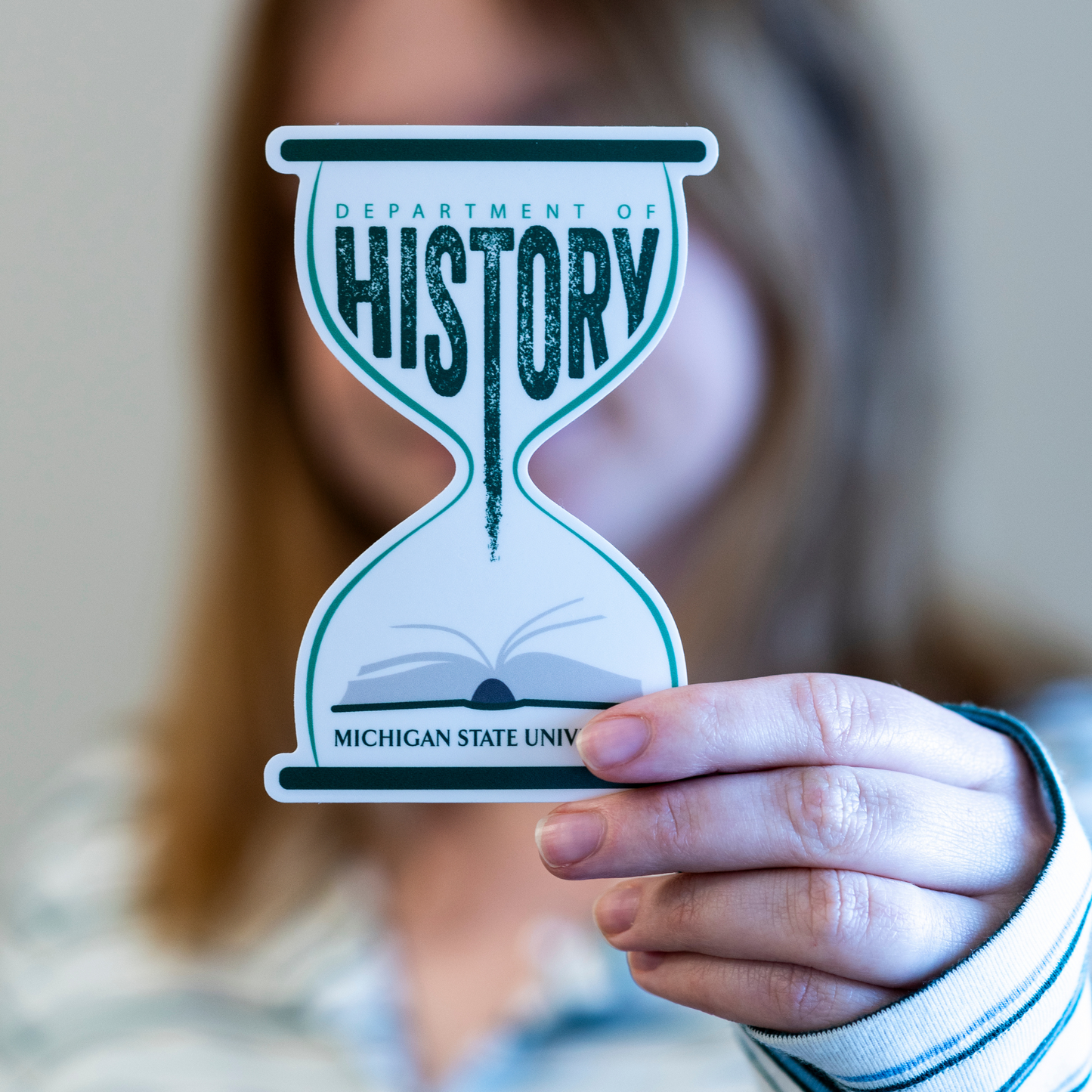 Person holding a forest green and white vinyl sticker that features an hourglass shape, history book, and the text “Department of History”. 