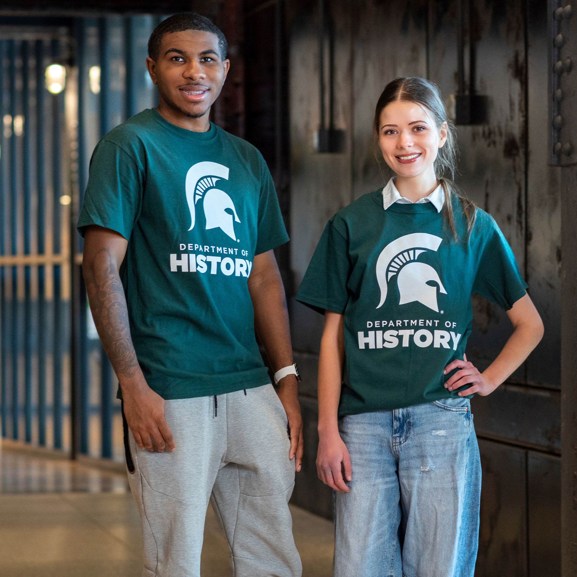 Two individuals wearing forest green T-shirts featuring a Spartan Helmet logo above the text "Department of History" printed in white.