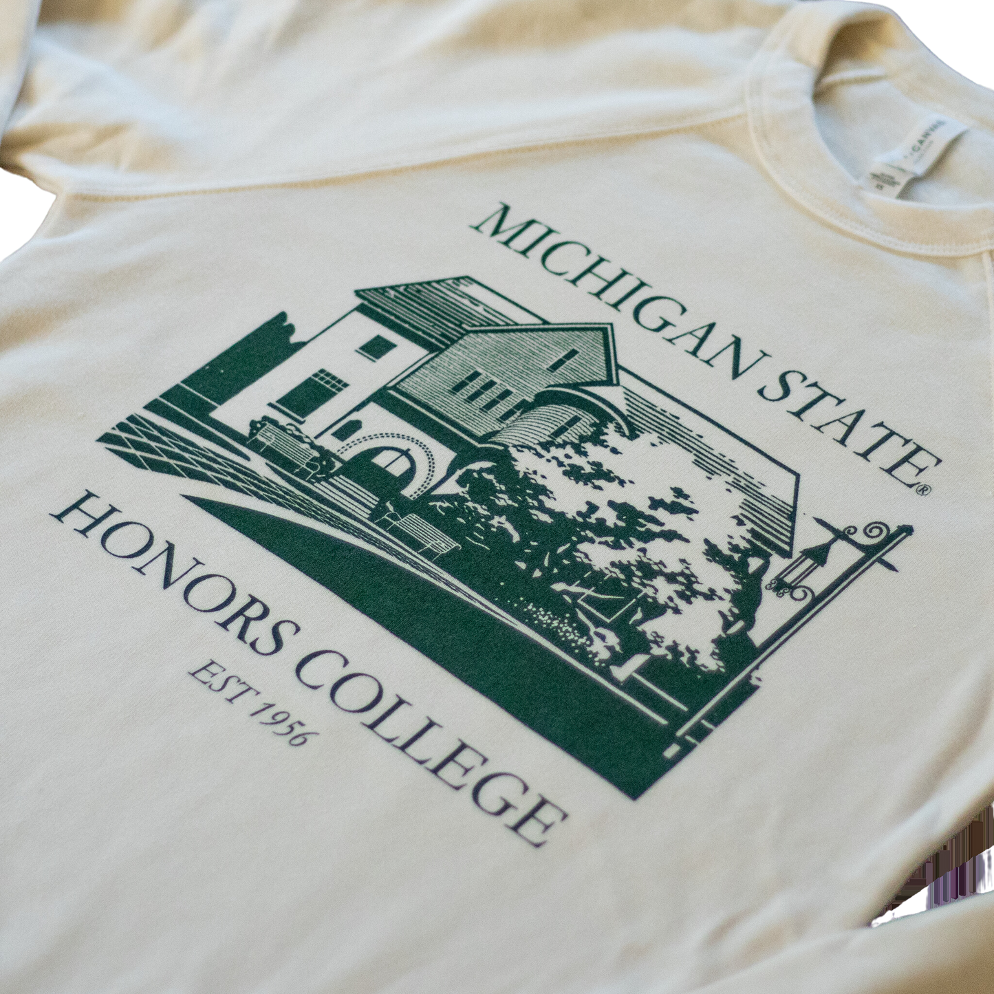 Close up of a natural/tan colored crewneck sweatshirt. On the torso of the sweatshirt in green is an image of the first MSU honors college building. Around the image is the text "Michigan State Honors College est. 1956."