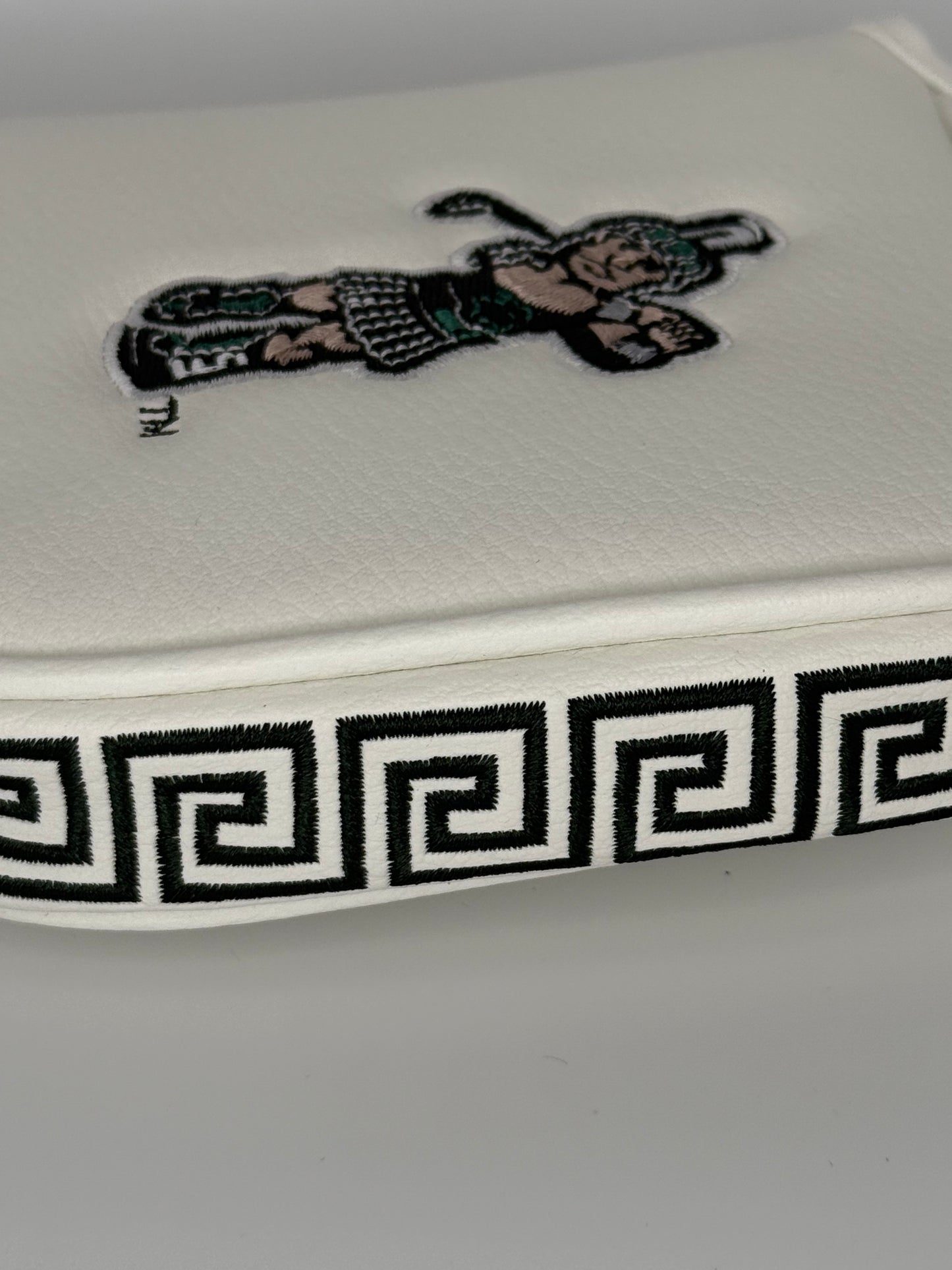 Close up of a white mallet putter headcover with an embroidered Golf Sparty logo on the front and a green and white Greek design on the side. 