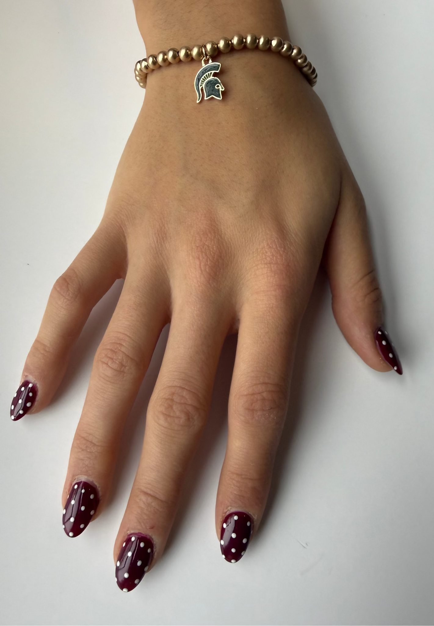 Hand with gold color beads with a small green and gold Spartan Helmet logo charm dangling down. 