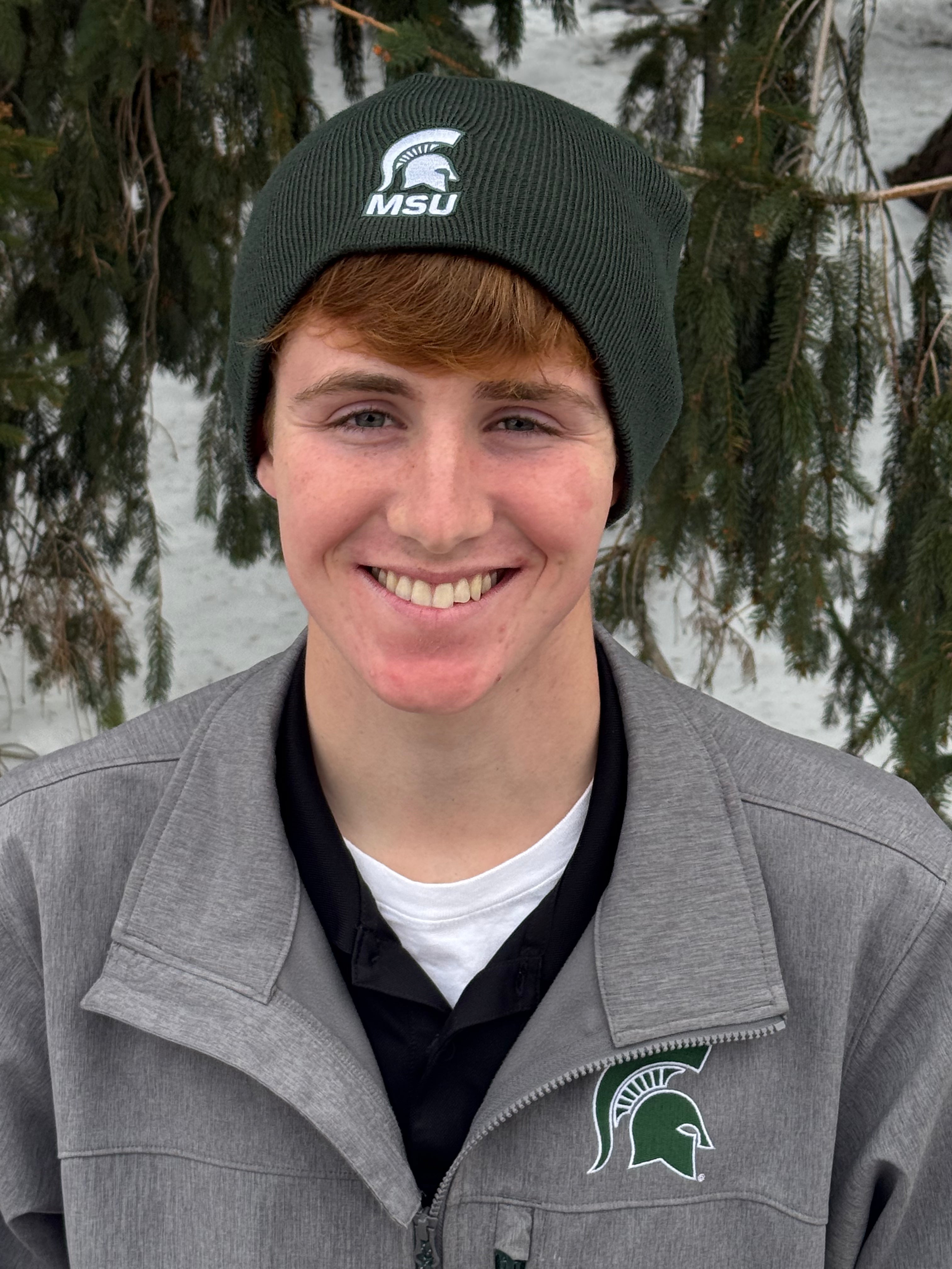 Man standing outdoors, wearing a dark green knit beanie with a Spartan Helmet logo embroidered on the bottom section and below that is an embroidered wordmark that states "MSU."
