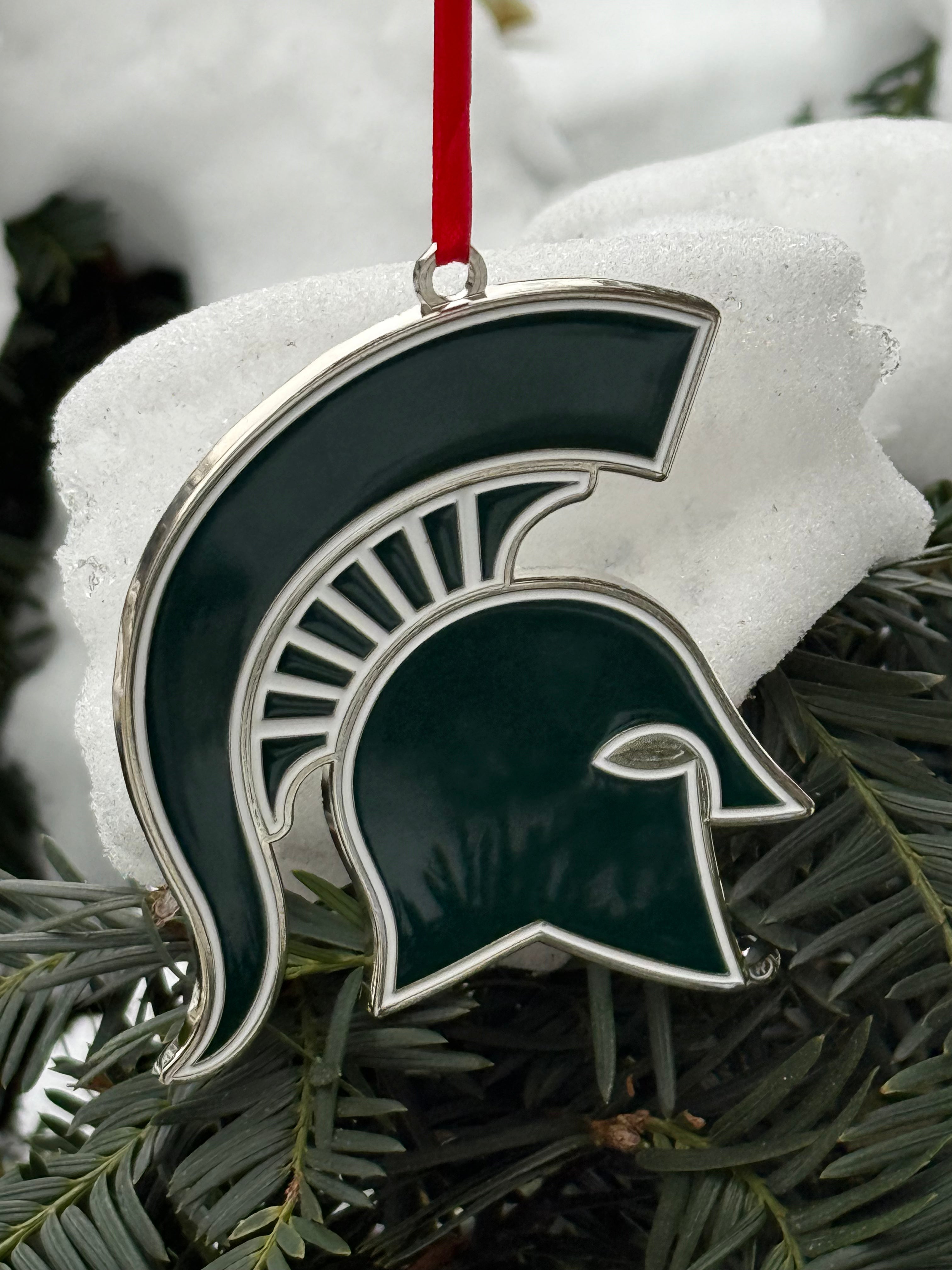 Metal ornament shaped as a Spartan helmet logo. The ornament is dark green with a silver outline and has a red ribbon attached to it. 