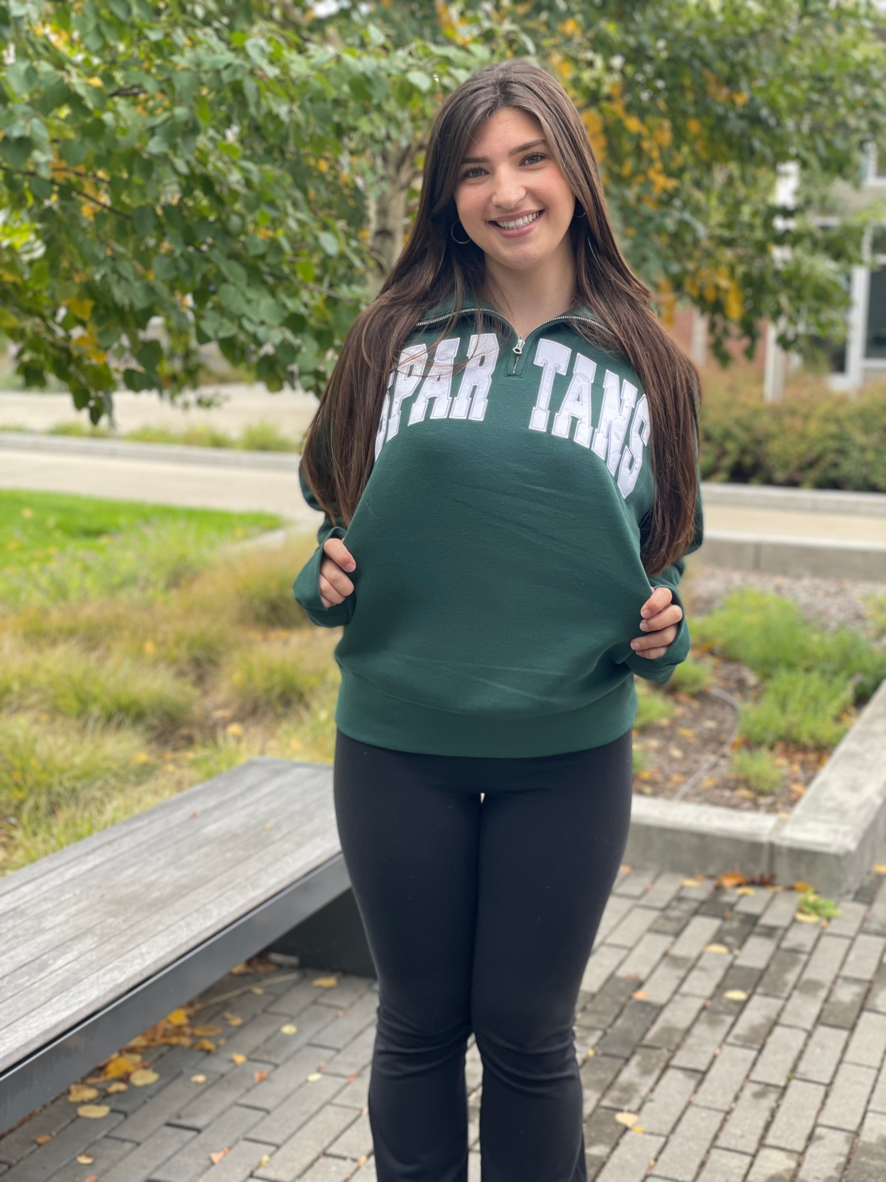 Person wearing a green quarter-zip sweatshirt with 'SPARTAN' in white satin applique letters on the upper chest. 