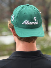 Dark green baseball cap. On the front panels in white cursive embroidery, is "Alumni" with a white Spartan Helmet logo above the letter n. On the side of the hat is a white block S. 