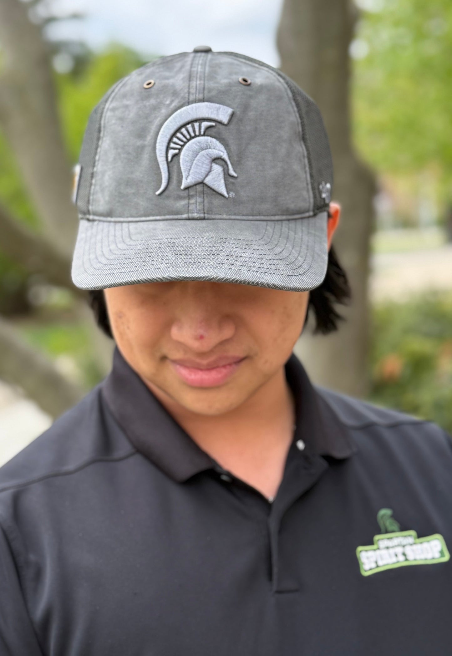 Person standing outdoors showing a gray hat with a light gray Spartan Helmet logo embroidered on the front. The sides of the hat are gray mesh. 