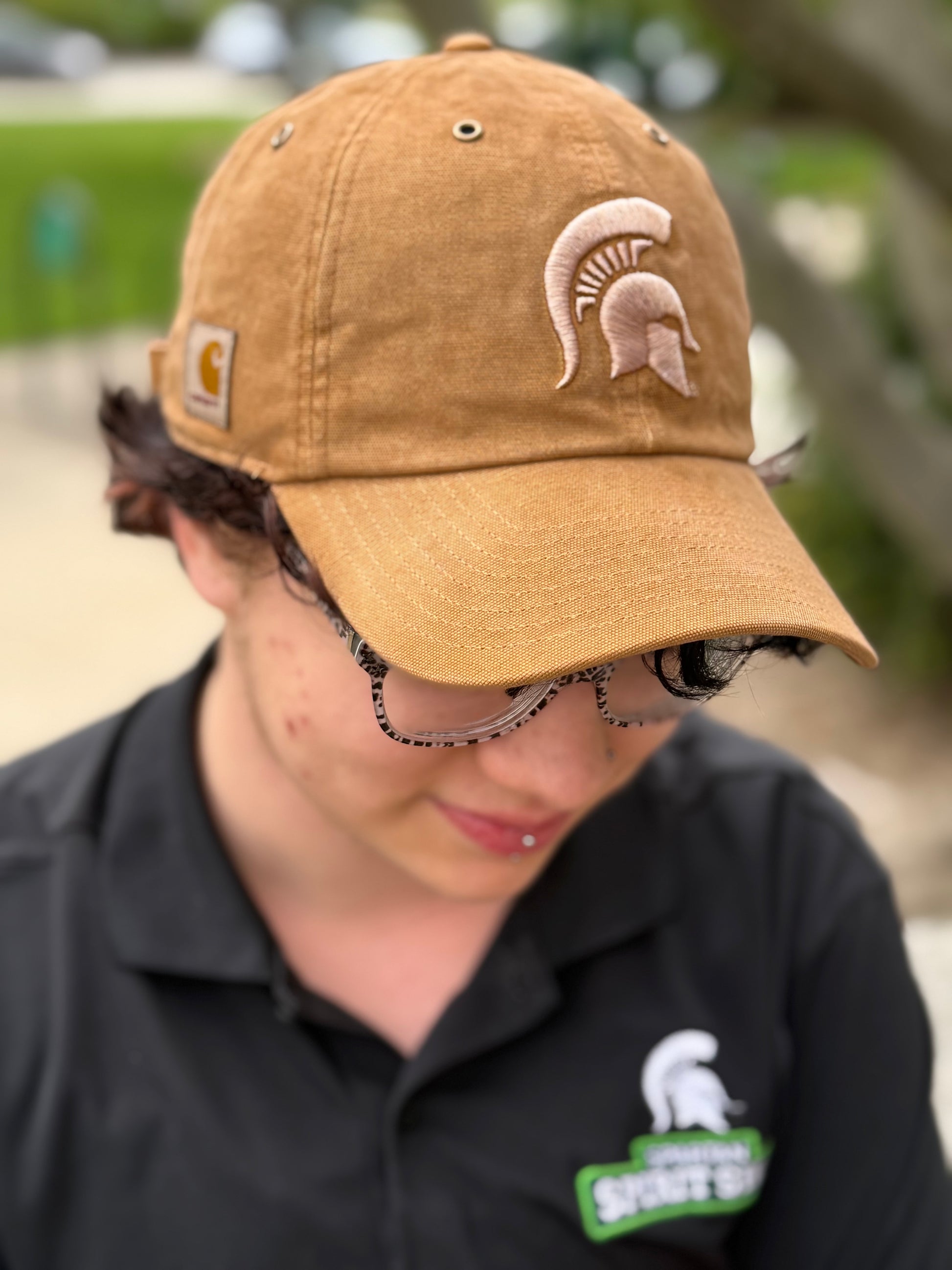 Person standing outdoors showing a beige hat with a light beige Spartan Helmet logo embroidered on the front and Carhartt logo patch on the right side. 